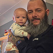 Jameson is registered to the contest to win money with this photo: baby, man, beard, tattoo, watch, indoor, holding, pajamas, dinosaur, face, person, portrait, curious, clothing, hand, calendar, room, adult, child, casual