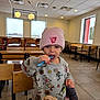 toddler, child, beanie, sweater, spoon, drink, table, restaurant, indoor, wooden_chair, food, snack, cup, window, floor, ceiling, light, person, eating, cute