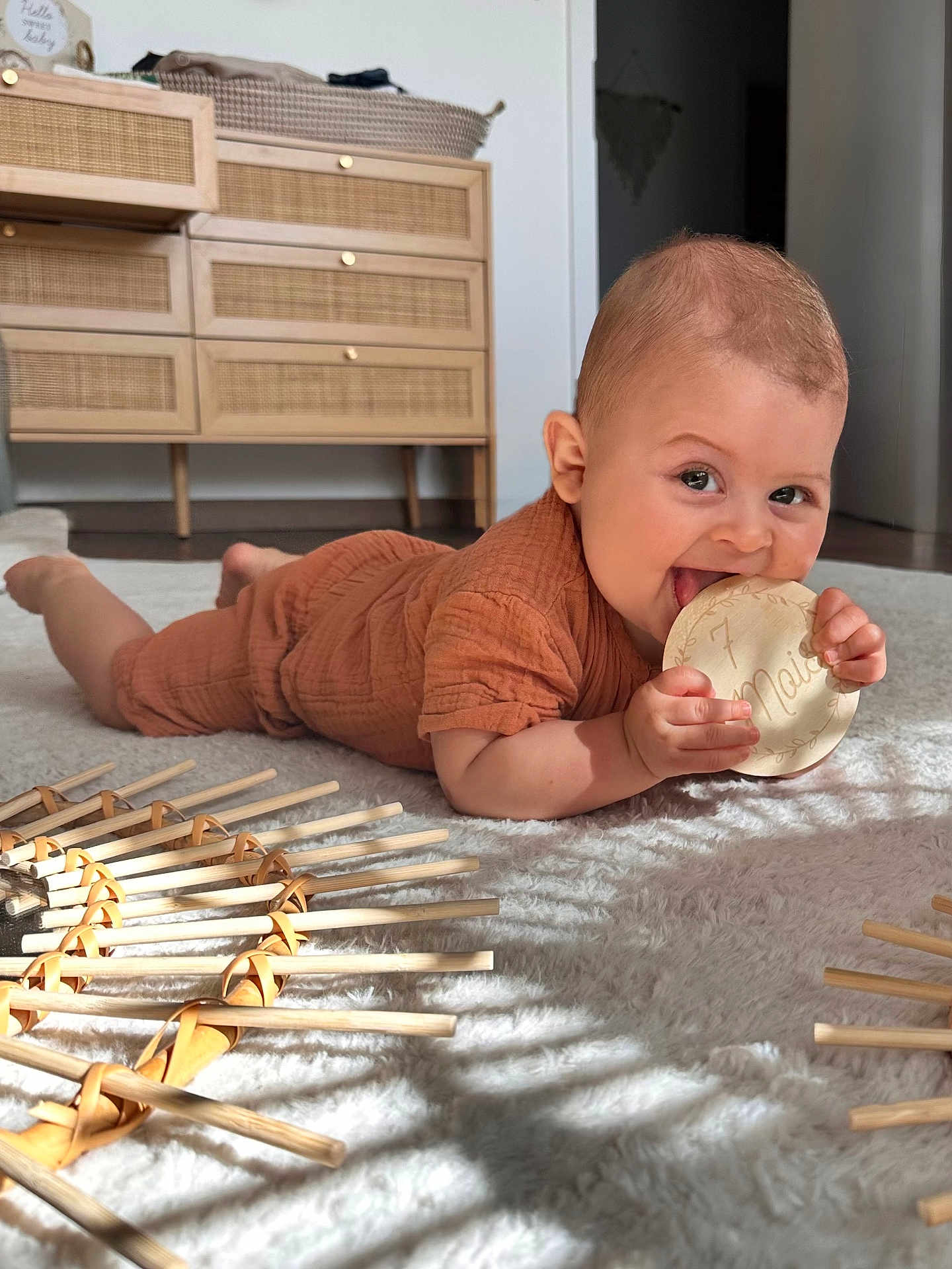 Andréa participe au concours pour gagner de l'argent avec cette photo : baby, infant, child, carpet, floor, wooden_toy, milestone_marker, orange_outfit, smiling, chewing, indoor, natural_light, wooden_furniture, cabinet, decor, happy, crawling, person, cozy, home