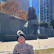 Thomas is registered to the contest to win money with this photo: toddler, child, baby, hat, beanie, pom_pom, jacket, fuzzy_clothing, grass, autumn, leaves, urban, city, buildings, water_feature, sunlight, outdoor, park, sitting, fall