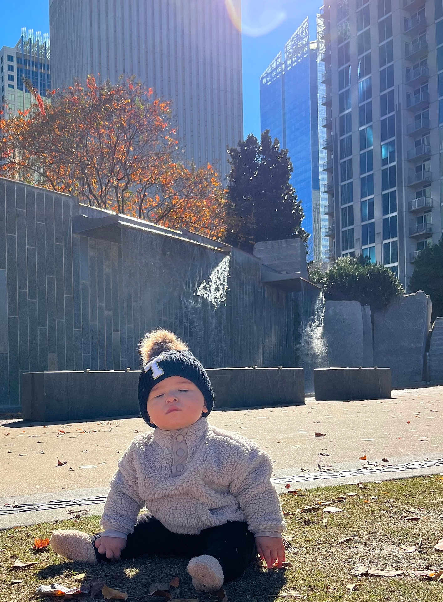 Thomas is registered to the contest to win money with this photo: toddler, child, baby, hat, beanie, pom_pom, jacket, fuzzy_clothing, grass, autumn, leaves, urban, city, buildings, water_feature, sunlight, outdoor, park, sitting, fall