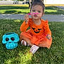 toddler, child, grass, outdoor, orange_dress, halloween, skull_bucket, bubble_wand, playful, smiling, cute, sitting, daylight, suburban, tree, sidewalk, shoes, greenery, fun, holiday