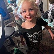 Lainey is registered to the contest to win money with this photo: child, girl, smile, blonde_hair, headband, flower, black_shirt, text_on_shirt, indoor, carpet, furniture, table, other_children, barefoot, playful, messy_room, curtain, toy, person, happy