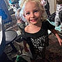 child, girl, smile, blonde_hair, headband, flower, black_shirt, text_on_shirt, indoor, carpet, furniture, table, other_children, barefoot, playful, messy_room, curtain, toy, person, happy