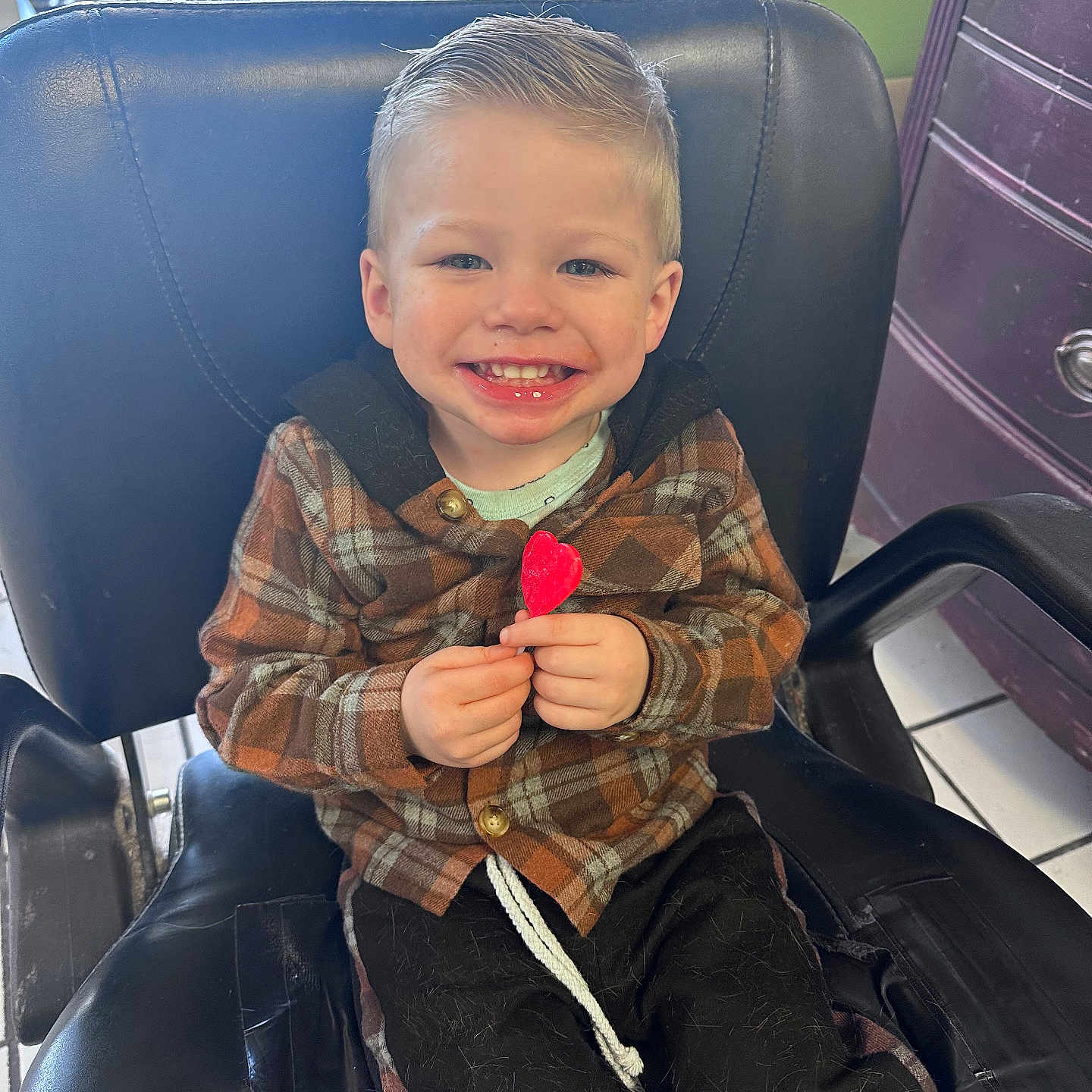 Zachariah is registered to the contest to win money with this photo: black_chair, boy, cabinet, chair, child, clothing, cute, floor_tiles, hair, hands, happy, heart_shaped, indoor, light, lollipop, plaid_jacket, portrait, room, seated, smiling