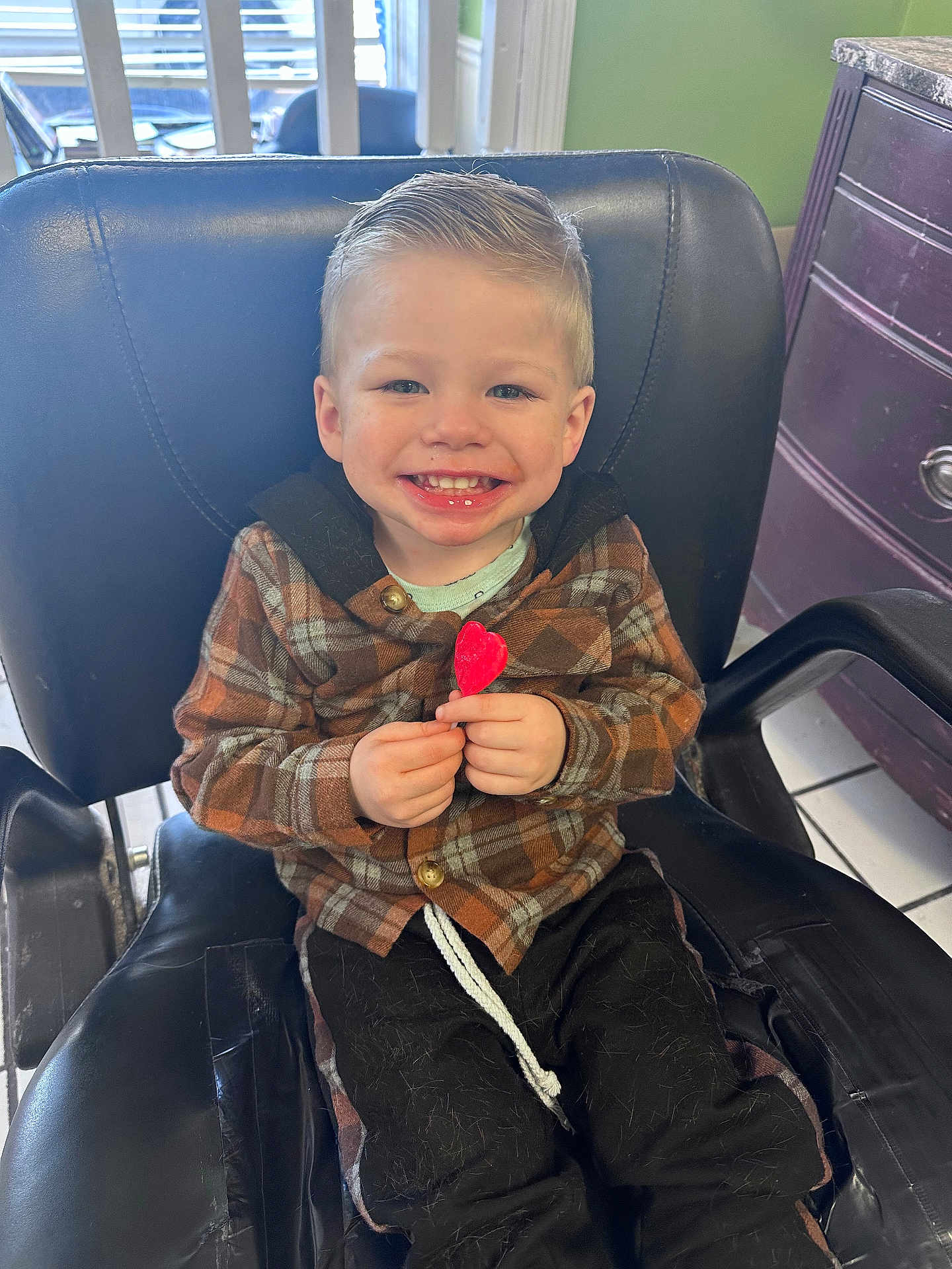 Zachariah is registered to the contest to win money with this photo: child, boy, smiling, plaid_jacket, lollipop, heart_shaped, chair, indoor, black_chair, hair, happy, cute, portrait, seated, hands, clothing, floor_tiles, cabinet, room, light