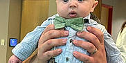 Ledger is registered to the contest to win money with this photo: baby, child, person, hands, bow_tie, shirt, pants, watch, ring, indoor, exit_sign, surprised_expression, adult_hands, holding, close_up, portrait, clothing, face, skin, furniture
