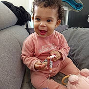 Kaïly participe au concours pour gagner de l'argent avec cette photo : baby, child, curly_hair, pink_outfit, pacifier, beads, toy, couch, grey, smiling, happy, indoors, plush_toy, seated, cute, infant, clothing, holding, home, playful