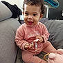 baby, child, curly_hair, pink_outfit, pacifier, beads, toy, couch, grey, smiling, happy, indoors, plush_toy, seated, cute, infant, clothing, holding, home, playful