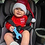 baby, car_seat, child, smiling, red_clothing, santa_hat, toy, blue_toy, winter_clothing, cozy_booties, face, person, seatbelt, indoor, car_interior, happy, holiday, festive, cute, infant
