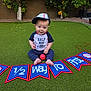 Everhett joined the competition — help win amazing prizes! baby, child, baseball, cap, grass, outdoor, toy, ball, banner, celebration, milestone, half_birthday, sitting, smiling, blue_clothing, red_ball, hat, backyard, greenery, cute