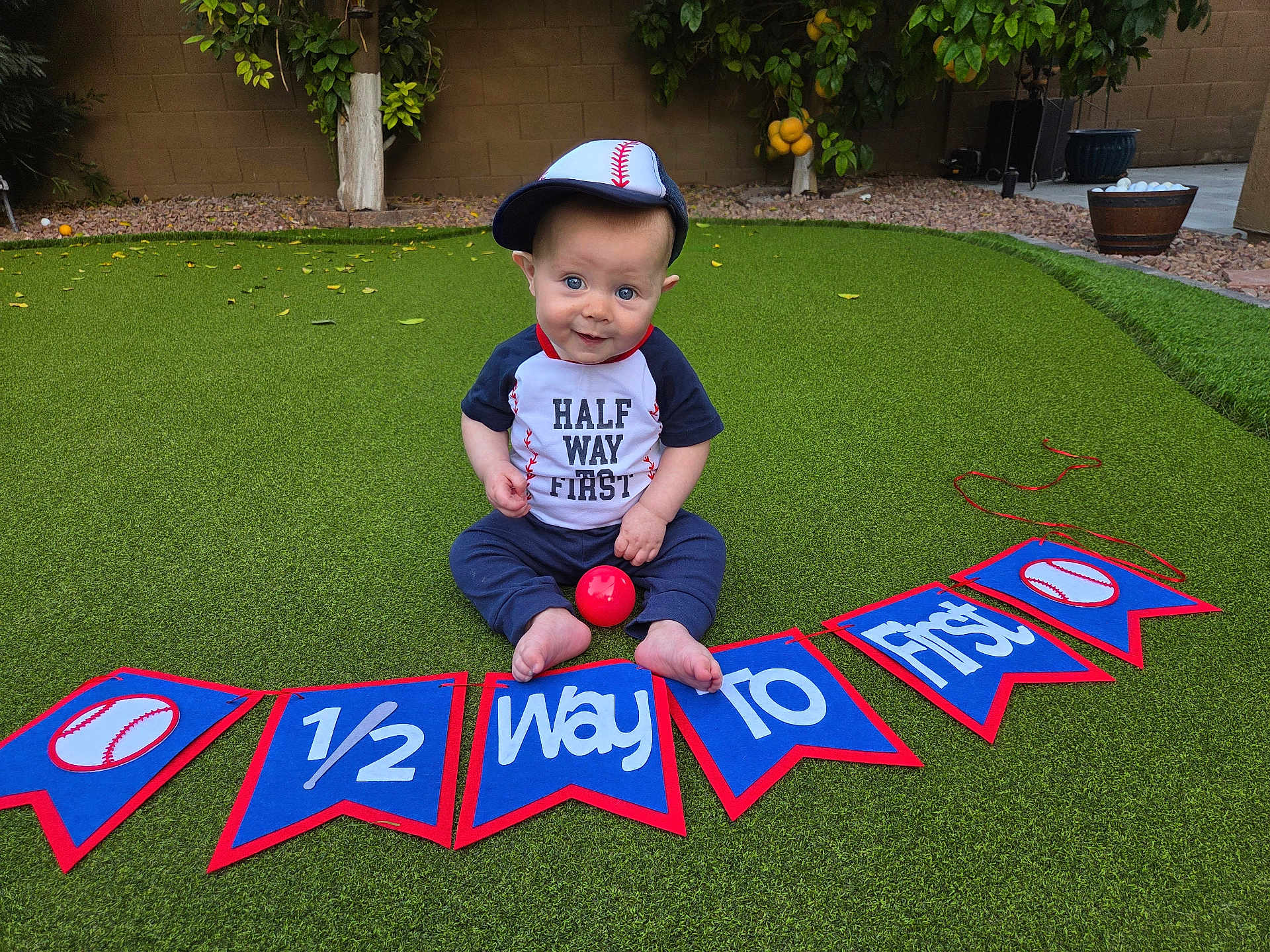 Everhett joined the competition — help win amazing prizes! baby, child, baseball, cap, grass, outdoor, toy, ball, banner, celebration, milestone, half_birthday, sitting, smiling, blue_clothing, red_ball, hat, backyard, greenery, cute