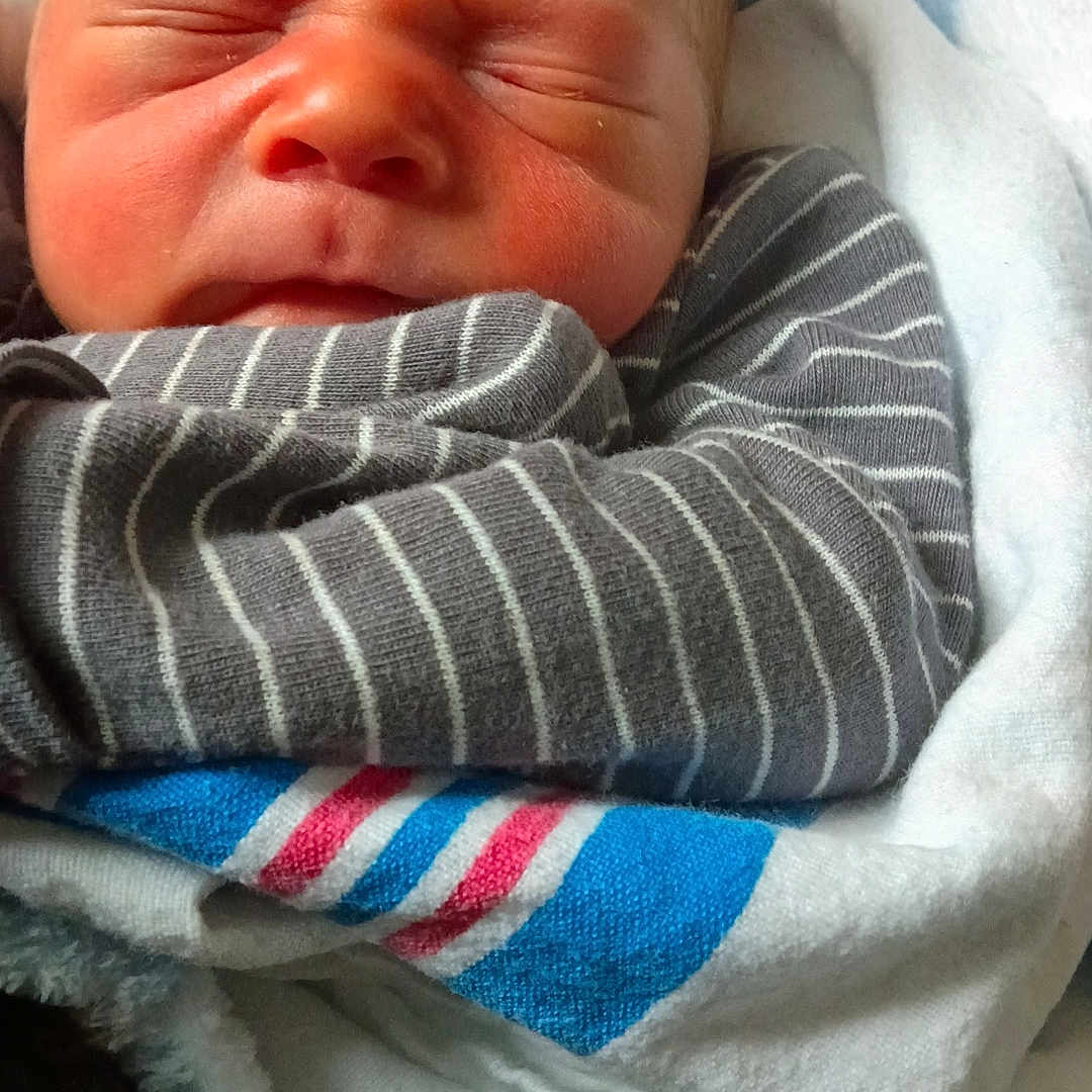 Leeland is registered to the contest to win money with this photo: newborn, baby, infant, child, sleeping, scrunching, face, portrait, bundled, blanket, swaddled, striped_clothing, gray, blue, white, red, cozy, indoors, close_up, photography