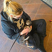 Atlanta is registered to the contest to win money with this photo: black_pants, brown_tiled_floor, cuddle, dog, glasses, holding, indoor, jacket, leash, looking_up, paws, person, ponytail, puppy, rug, sitting, slippers, small_dog, sweater, tile_floor