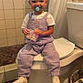 Addisynn is registered to the contest to win money with this photo: toddler, child, pacifier, bathroom, toilet, overalls, sneakers, white_shirt, tile_wall, trash_bin, floor_mat, curtain, smiling, person, indoor, holding_object, baby, cute, seated, plush