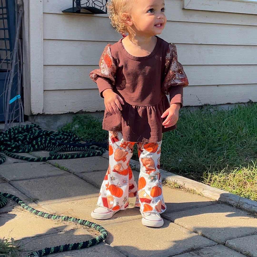 Addisynn is registered to the contest to win money with this photo: brown_top, child, curly_hair, daytime, footwear, garden_hose, grass, house_wall, long_sleeves, outdoor, pants, pavement, playful_clothing, pumpkin_pattern, shadow, smile, standing, sunlight, toddler, window