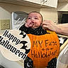 baby, pumpkin, halloween, costume, kitchen, cabinet, counter, hand, black_clothing, stars, moon, face, infant, celebration, holiday, text_overlay, bat, orange, person, cute