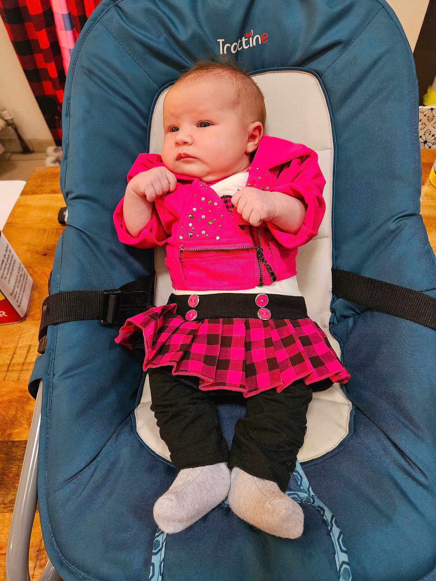 Elena a rejoint le concours — aidez-le/la à gagner de superbes lots ! baby, infant, child, pink_jacket, plaid_skirt, black_leggings, gray_socks, baby_seat, blue_cushion, indoor, wooden_table, curious_expression, clothing, seated, small_hands, cute, young_child, face, person, portrait