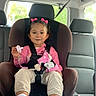 toddler, child, car_seat, seatbelt, pink_bows, pacifier, plush_toy, minnie_mouse, pink_shirt, beige_pants, white_sneakers, car_interior, window, headrest, safety, smiling, cute, young_child, hair_bows, child_safety