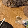 cat, sleeping, tabby, placemat, tablecloth, golden, pattern, black_plate, rice, spoon, beads, craft_supplies, plastic_bag, table, indoor, pet, resting, close_up, feline, cozy