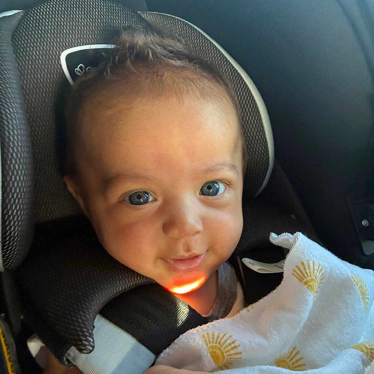 Sebastian is registered to the contest to win money with this photo: baby, child, car_seat, blanket, sunlight, smile, face, hand, seatbelt, portrait, infant, person, indoors, cute, cozy, clothing, headrest, bright_eyes, happy, young