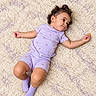 baby, child, infant, portrait, lying_down, smiling, happy, curly_hair, purple_clothing, romper, socks, rug, carpet, indoor, overhead_view, close_up, arms_outstretched, holding_object, soft_lighting, adorable