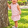 child, toddler, girl, smiling, curly_hair, hair_bow, pink_outfit, dress, flower, tree, park, playground, grass, outdoors, summer, portrait, standing, shoes, happy, sunlight