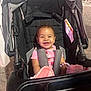 baby, stroller, smiling, pink_clothing, blanket, bracelet, child, happy, portrait, seatbelt, outdoor, curly_hair, person, infant, toy, black_stroller, cute, face, sitting, night