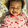 baby, child, smiling, tongue_out, onesie, cherry_pattern, pink_clothing, curly_hair, portrait, close_up, indoors, floral_background, cute, happy, infant, person, face, playful, cozy, sleepwear
