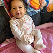 Kaia is registered to the contest to win money with this photo: baby, infant, child, smiling, happy, portrait, indoors, onesie, pajamas, pink, white, blanket, bed, sitting, face, eyes, hair, couch, patterned_background, home