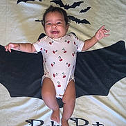 Kaia is registered to the contest to win money with this photo: baby, child, onesie, cherry_pattern, blanket, bat, black_bat, smile, happy, infant, lying_down, cute, text, outstretched_arms, baby_bat_text, portrait, indoor, soft_texture, head, feet