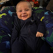 Masen is registered to the contest to win money with this photo: baby, smiling, child, onesie, blue_clothing, dinosaur_pattern, cushion, indoor, flowers, homedecor, happy, portrait, infant, seated, cozy, cute, person, face, young, comfortable