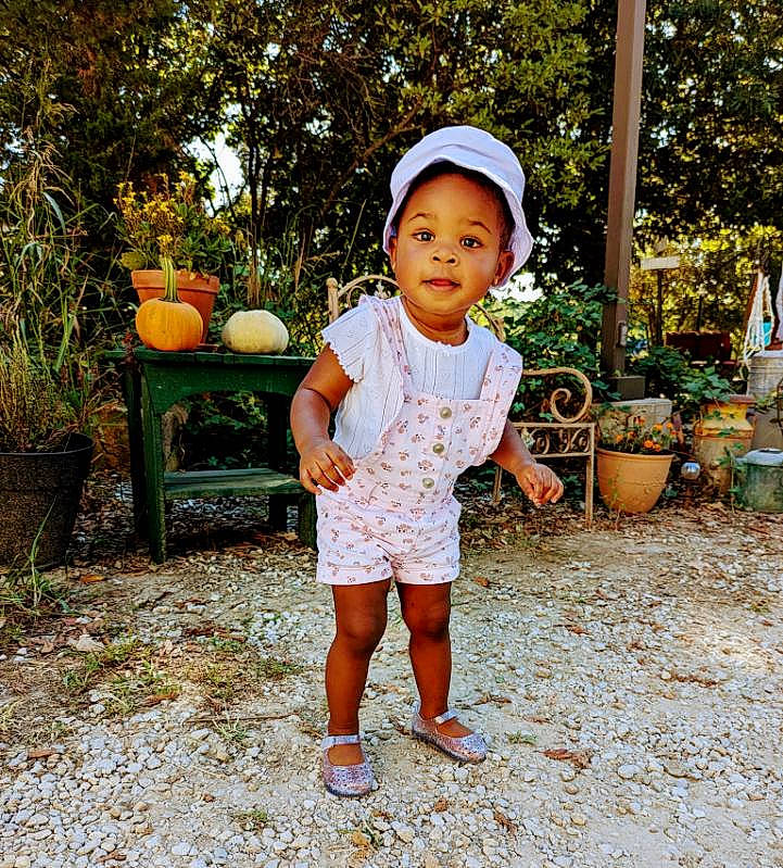 Zariyah is registered to the contest to win money with this photo: baby_toddler_clothing, child, flowerpot, fun, grass, happy, hat, houseplant, leaf, leisure, orange, people_in_nature, person, plant, shorts, sitting, sleeve, smile, t_shirt, toddler