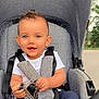 baby, blue_pants, child, clothing, daylight, face, gray, hair, hand, happy, keys, nature, outdoor, park, person, seatbelt, smile, stroller, toddler, white_shirt