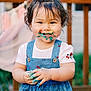toddler, child, paint, colorful, face_paint, blue_dress, cherry_pattern, smiling, happy, messy, hands, outdoor, wooden_fence, garland, celebration, curly_hair, white_shirt, portrait, daylight, cute