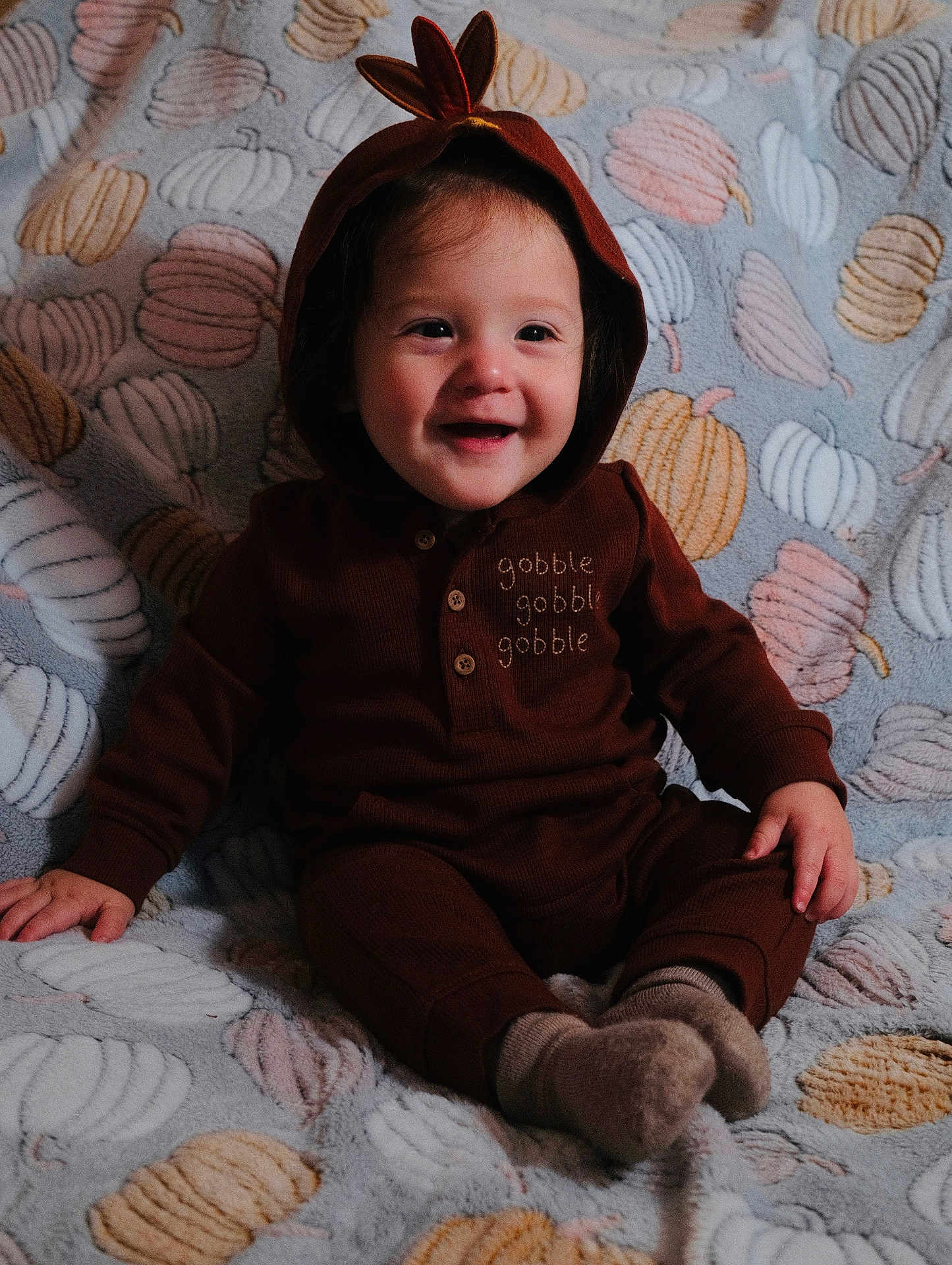 August is registered to the contest to win money with this photo: baby, child, smiling, hoodie, brown_clothing, sitting, blanket, pumpkin_pattern, cozy, indoors, cute, happy, face, portrait, infant, soft_texture, warm_clothing, toddler, playful, young_child
