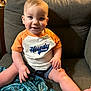 toddler, child, smiling, orange_shirt, shorts, couch, indoor, face, person, blue_eyes, baby, happy, sitting, hand, legs, blanket, fabric, casual_clothing, cute, home