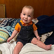 Charles joined the competition — help win amazing prizes! baby, child, smile, orange_shirt, denim_overalls, bed, pillow, blanket, wood_paneling, indoor, cute, happy, sitting, foot, hand, infant, person, cozy, portrait, casual