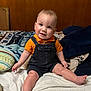 baby, child, smile, orange_shirt, denim_overalls, bed, pillow, blanket, wood_paneling, indoor, cute, happy, sitting, foot, hand, infant, person, cozy, portrait, casual