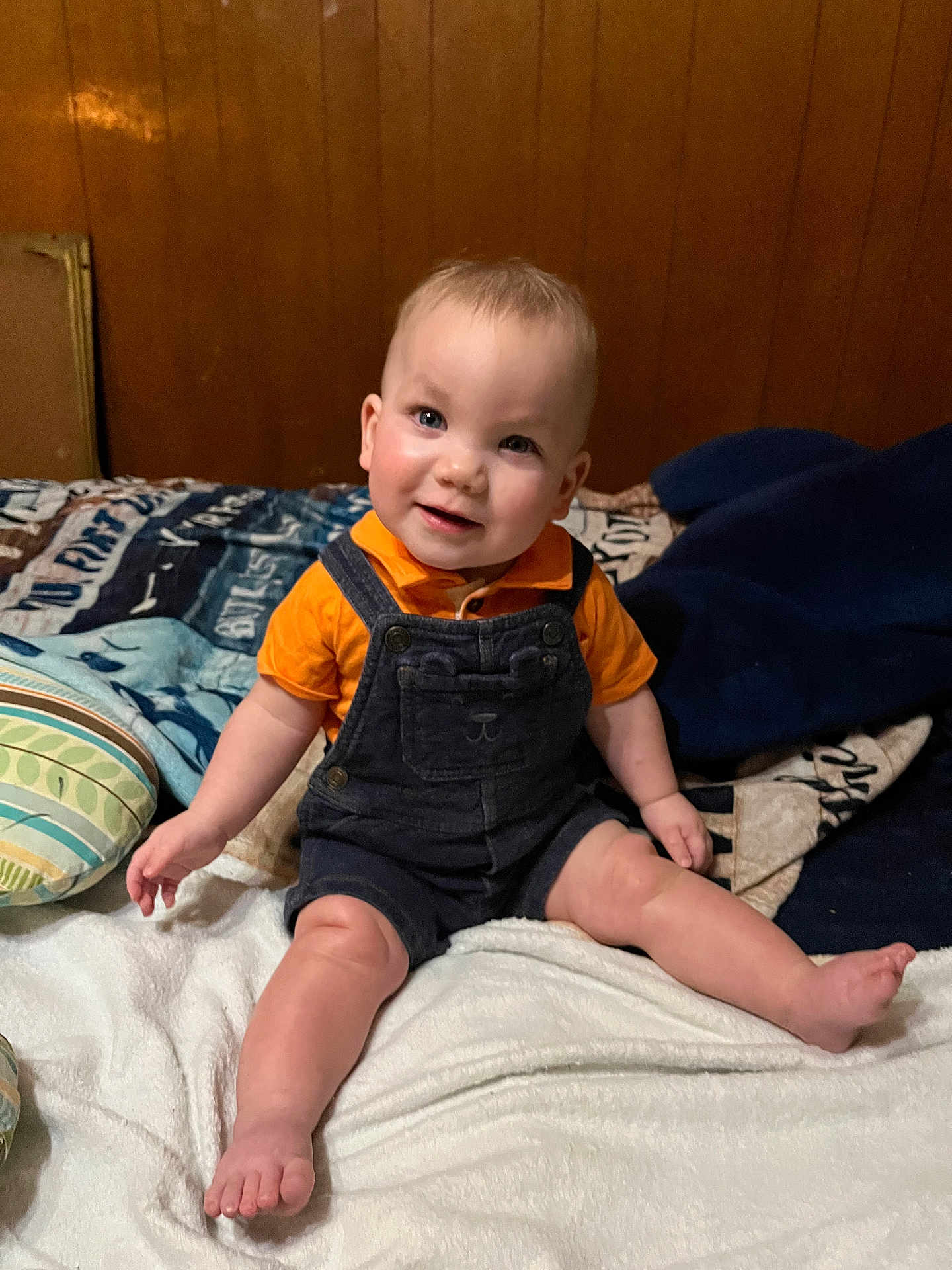 Charles joined the competition — help win amazing prizes! baby, child, smile, orange_shirt, denim_overalls, bed, pillow, blanket, wood_paneling, indoor, cute, happy, sitting, foot, hand, infant, person, cozy, portrait, casual