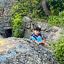 child, boy, rock, forest, nature, greenery, trees, outdoor, adventure, smiling, curious, daylight, backpack, plants, leaves, young, exploration, casual_clothing, natural_light, scenery