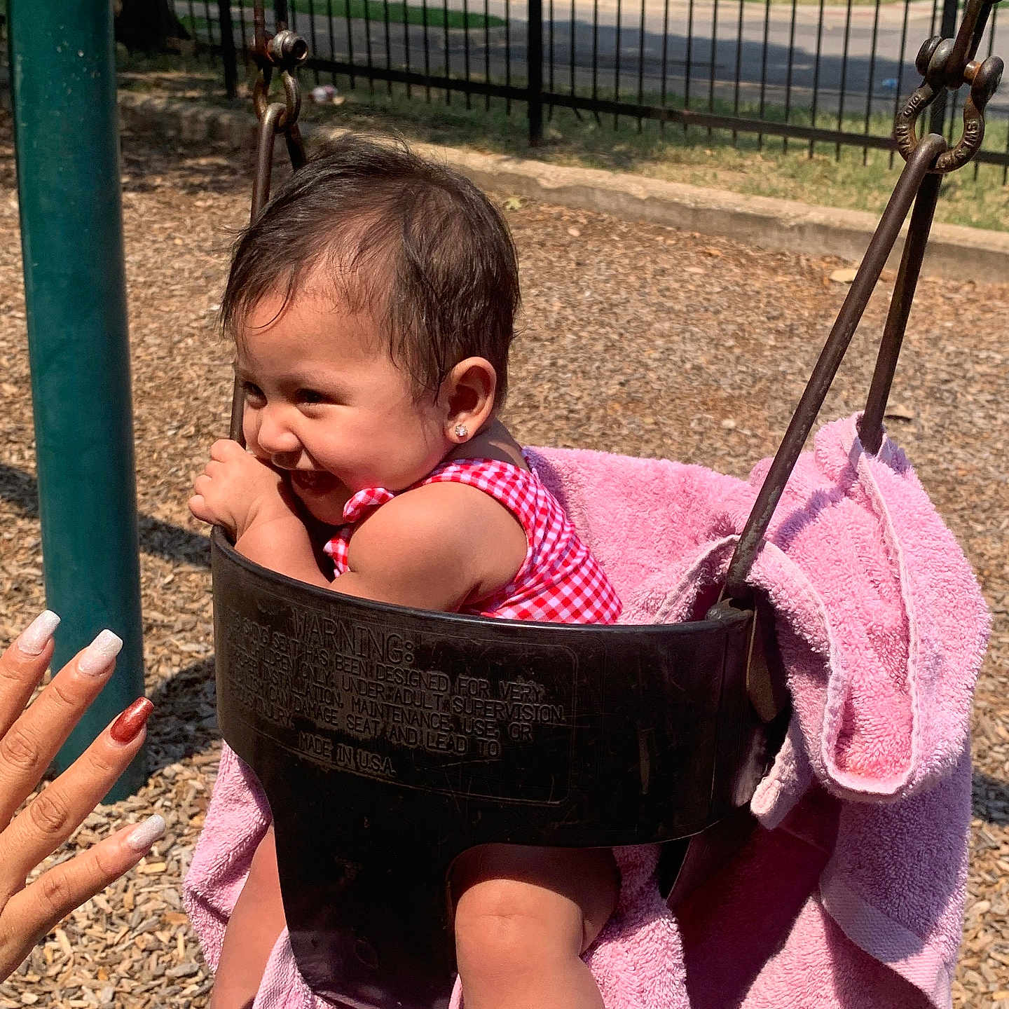 Gardenia is registered to the contest to win money with this photo: baby, child, earrings, fence, fun, hand, happy, metal_chains, outdoor, park, pink_towel, play, playground, red_clothing, smiling, summer, sunlight, swing, toddler, woodchips