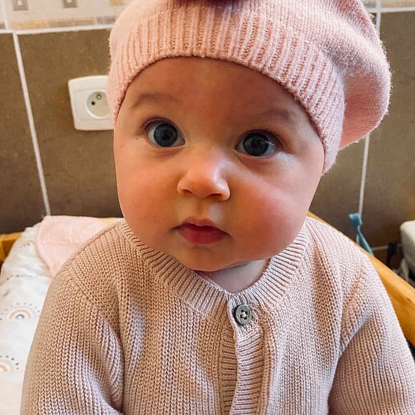 Yvanah a rejoint le concours — aidez-le/la à gagner de superbes lots ! baby, child, indoor, pink, knitwear, beret, flower, face, portrait, cute, wide_eyes, sitting, sweater, infant, headwear, expression, tile_wall, curious, buttoned, cozy