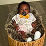 baby, infant, pacifier, bow_tie, white_clothing, basket, fur_lining, wooden_floor, indoors, cute, child, footwear, sitting, portrait, young_child, comfortable, cozy, smiling, person, adorable