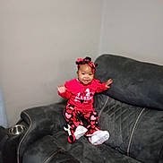 Esther is registered to the contest to win money with this photo: baby, infant, smiling, sitting, red_outfit, heart_pattern, headband, bow, white_shoes, sofa, couch, armrest, gray_wall, living_room, plush_toy, upholstery, portrait, happy, toddler_clothing, cup_holder