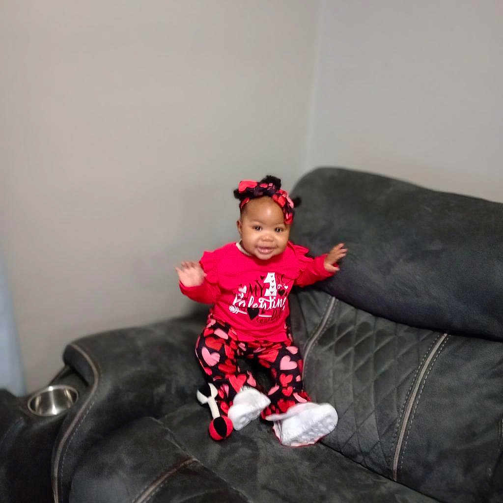 Esther is registered to the contest to win money with this photo: baby, infant, smiling, sitting, red_outfit, heart_pattern, headband, bow, white_shoes, sofa, couch, armrest, gray_wall, living_room, plush_toy, upholstery, portrait, happy, toddler_clothing, cup_holder