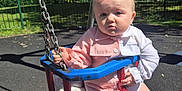Harper-Grace is registered to the contest to win money with this photo: baby, child, swing, playground, outdoor, park, sunny, trees, fence, clothing, white_socks, curious, puzzled_expression, seat, metal_chains, red, blue, person, infant, daytime