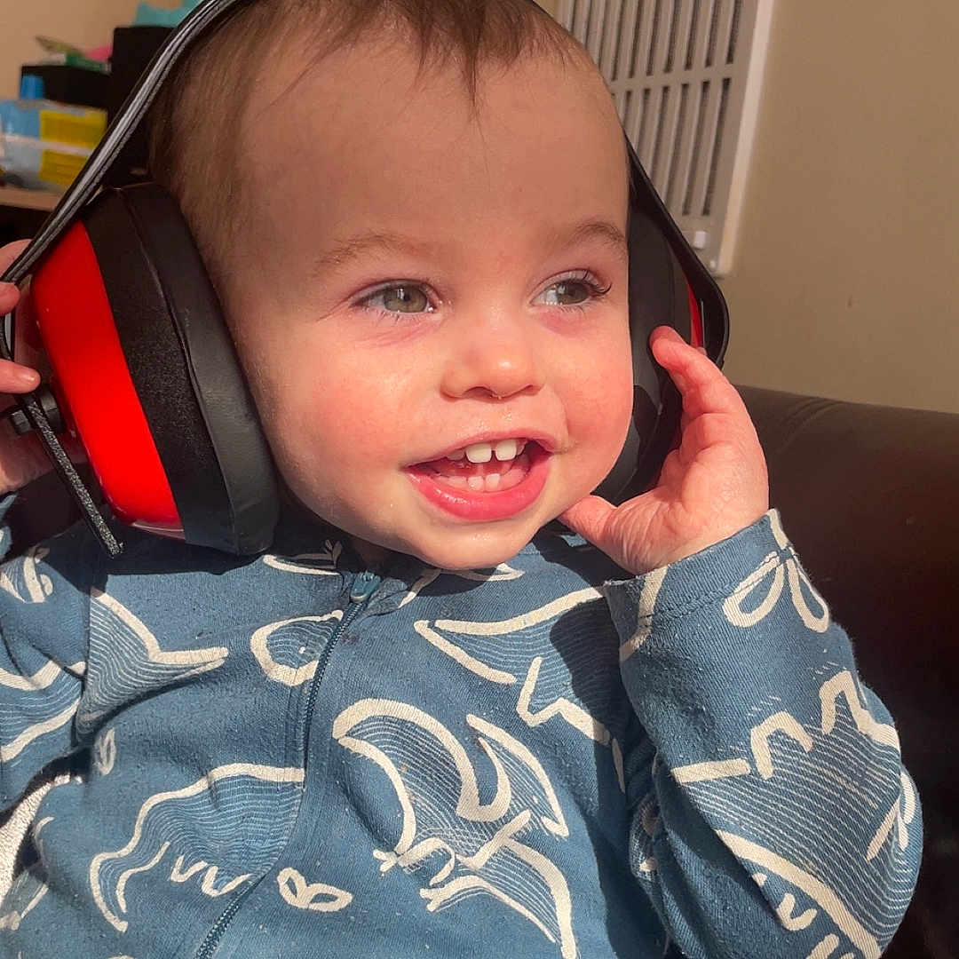 Arlo-George joined the competition — help win amazing prizes! blue_clothing, child, couch, cozy, cute, dinosaur_pattern, ear_protection, face, hair, hands, happy, headphones, indoor, infant, person, playful, smiling, sunlight, teeth, toddler