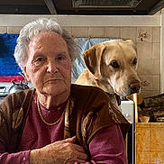 Edgar participe au concours pour gagner de l'argent avec cette photo : elderly_woman, dog, golden_retriever, kitchen, table, indoor, portrait, seated, warm_clothing, gray_hair, chain_necklace, tiles, teapot, wood_logs, appliance, cupboard, serious_expression, domestic, companionship, pet