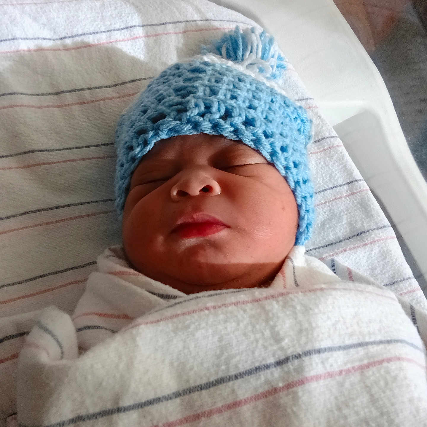 Hassan is registered to the contest to win money with this photo: newborn, baby, infant, sleeping, knitted_hat, blue_hat, swaddled, blanket, hospital, bassinet, portrait, face, closed_eyes, peaceful, indoors, stripes, textile, bedding, nursery, photography